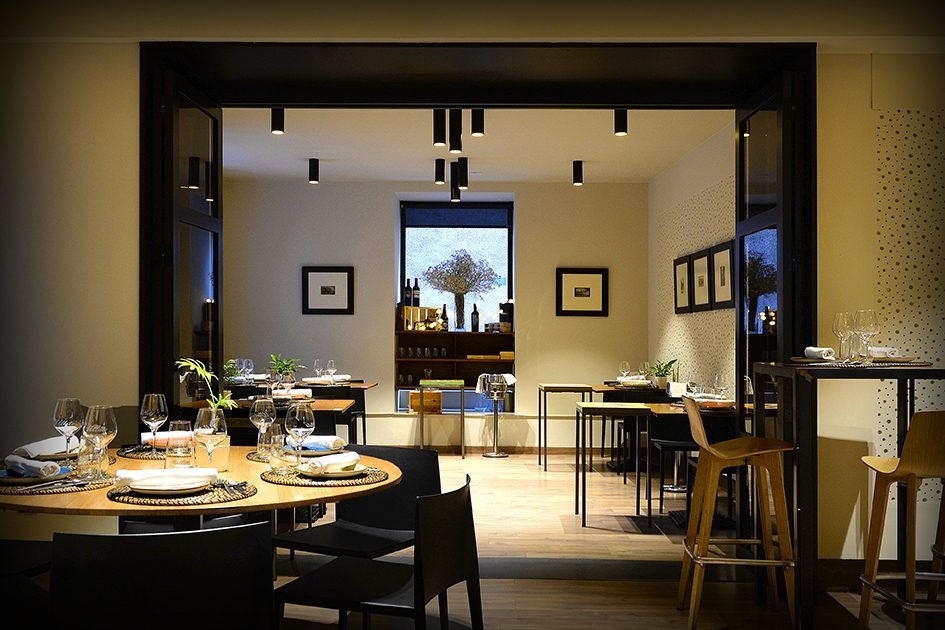 El comedor de Ullate: mesas de madera puestas, estantería de vinos al fondo y una ventana con un olivo